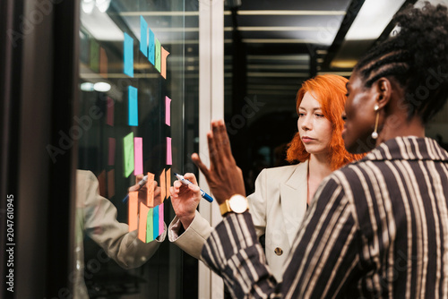 Two colleagues brainstorm at a glass wall in a modern office, arranging colorful sticky notes and planning ideas during a collaborative team session.