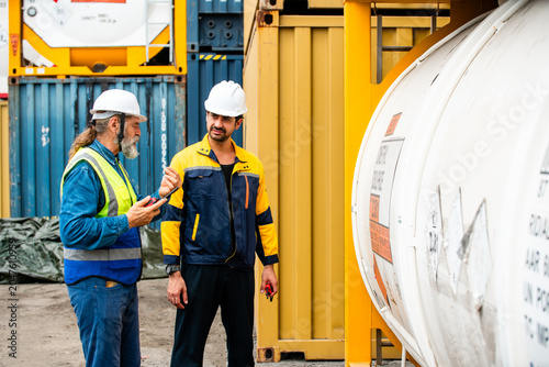 Two industrial workers in hard hats discuss an inspection beside a tank container at a shipping yard, surrounded by stacked cargo containers and equipment.