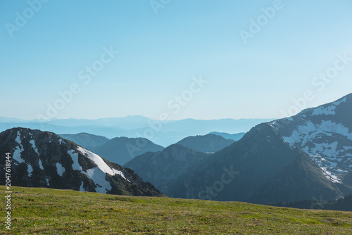 Scenic view from sunlit grassy meadow to abyss of alpine valley between big rocky mountains with snows and hilly layers of ridge silhouettes far away under clear blue sky in bright sun in sunny day.