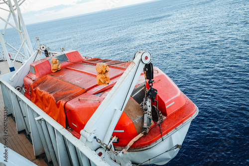 Schilderij op canvas High angle view of an orange enclosed emergency lifeboat marked for maximum 150 persons, mounted on a passenger vessel deck with the blue sea in the background
