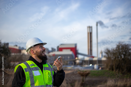 Engineer worker wearing safety vest and hard hat, looking up and using smartphone at industrial facility