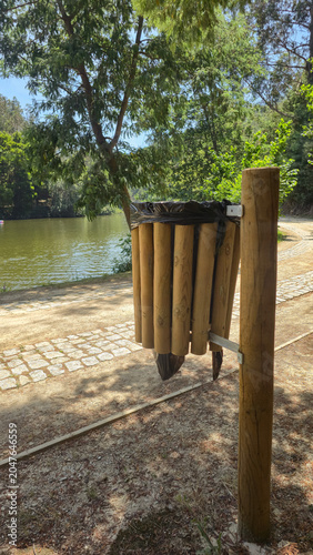A wooden trash bin along a lakeside walkway in a green park, next to a boat. Themes: environmental responsibility, public cleanliness, sustainable recreation, and nature conservation.