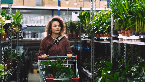 A happy woman with curly hair buys potted flowers at a garden center. The woman carries the flowers in a shopping cart.