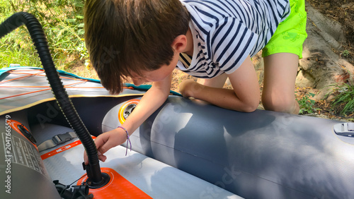 A child helps his father inflate an inflatable kayak with a hand pump on a forest trail near a river. Topics: outdoor adventures, active lifestyle, sustainable tourism, and water sports.