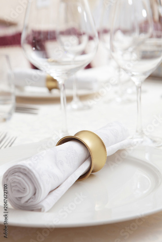 Setting festive table with white tablecloth. White serving napkin with yellow metal ring is placed on plate. Empty glasses for drinks. Concept of festive events. Preparation for the reception of