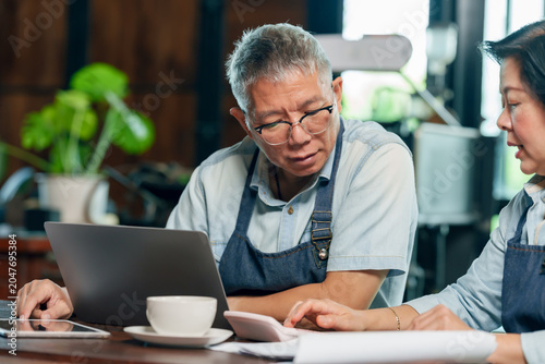Senior asian man cafe owner reviewing data on laptop while woman partner explaining business report teamwork concept small business management communication collaboration entrepreneurship lifestyle