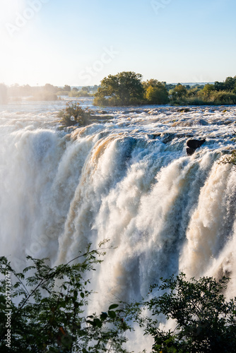 Fotografi Victoria Falls on the Zambia and Zimbabwe border shows white water plunging over a wide cliff, spray and mist rising, river churning above the brink in daylight