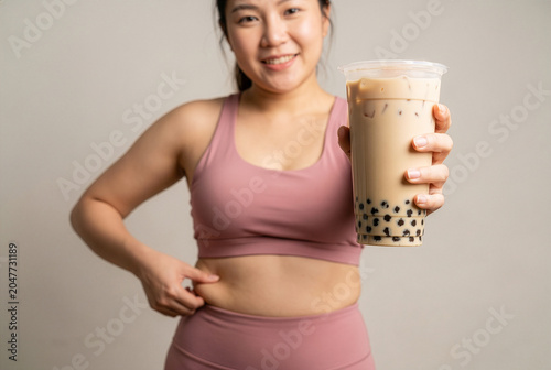Smiling Asian woman in sportswear holding bubble tea while pinching her belly fat, diet failure and cheat day concept