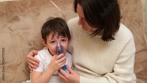 Woman helping kid use nebulizer inhalation mask for respiratory disease treatment at home. Child receiving steam for health.