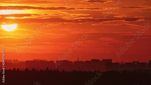 Sunset sky over city skyline with silhouette of trees, time lapse. Bright orange sun disk lowering behind clouds at dusk. Dramatic evening landscape view for weather background and travel concept.