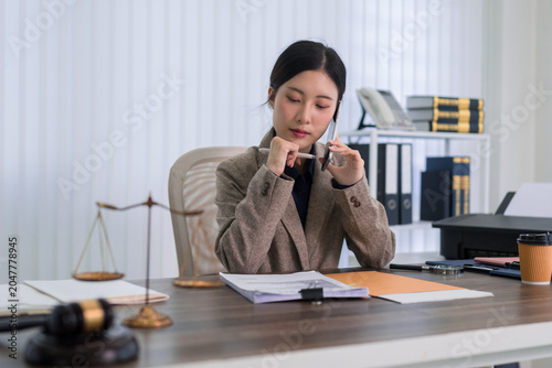 Asian female lawyer reviewing legal documents talking phone consultation professional office desk with scales justice gavel paperwork attorney workplace