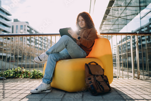 Fotografie Young professional with backpack sitting in yellow chair using digital tablet in modern city