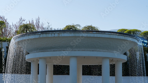 Krasnodar, Cloud Park. Circular rotunda with cascading water flowing down its sides like curtains. Rotunda reflect in lake of love. Surrounding greenery includes bushes and trees. Galitsky Park.