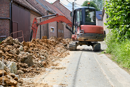 Mini excavator working on a narrow rural road, with a pile of excavated earth beside it. Construction and earthmoving in progress.