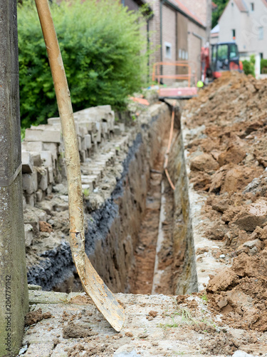A deep trench is dug for construction or utility work, with a shovel resting in the foreground. Piles of excavated earth line the site, and machinery is visible in the background, indicating ongoing b