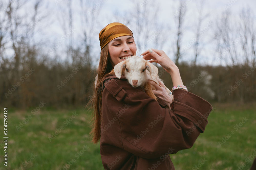 Naklejka premium A joyful woman in a brown coat warmly holds a white goat kid in an open field, conveying care and connection in a natural outdoor setting, a clear lifestyle moment with animal companionship.
