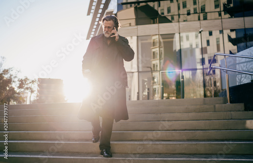 Foto Senior business leader walking downstairs toward sunlight while having mobile call near office, mature professional in trench coat holding coffee with lens flare and copy space background