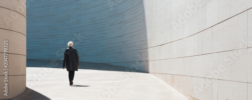 Elegant older woman active aging modern architecture minimal background senior walking urban curve white wall long shadow solitude daylight scene graceful senior walking along curved modern