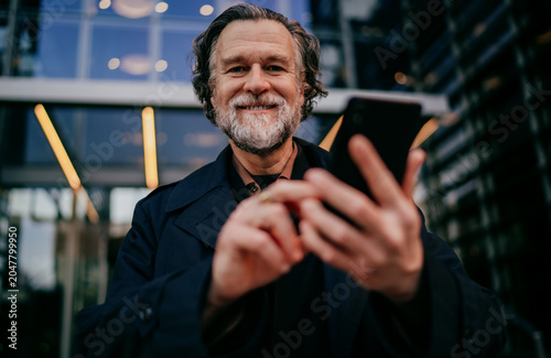 Portrait of cheerful senior businessman holding smartphone and looking at camera with joy, successful mature male professional using mobile device in contemporary city environment, copy space.