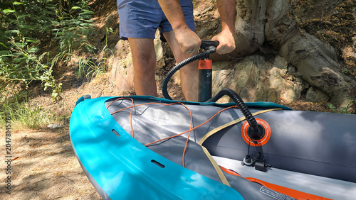 Person inflating an inflatable kayak with a hand pump on a forest trail near a river. Concepts of outdoor adventure, active lifestyle, sustainable tourism, and recreational water sports.