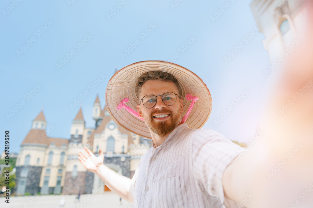 Fototapeta premium Caucasian young male tourist with Viet hat taking selfie in front of amusement park in Nha Trang, travel trip in Vietnam