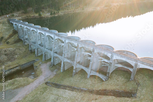 Elevated view of a concrete dam spanning a reservoir with warm evening sunlight reflecting on the water surface. Infrastructure and landscape concept
