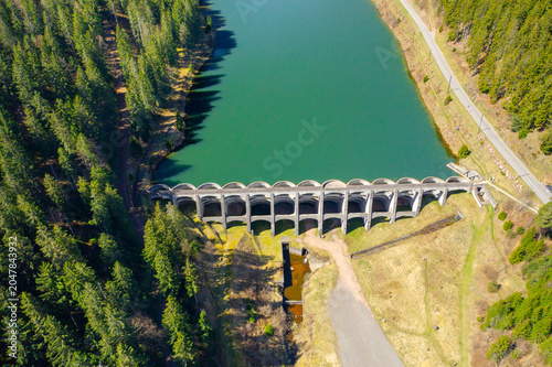Top-down drone photograph of a historic concrete dam holding a mountain reservoir surrounded by forest. Water resources and civil engineering concept
