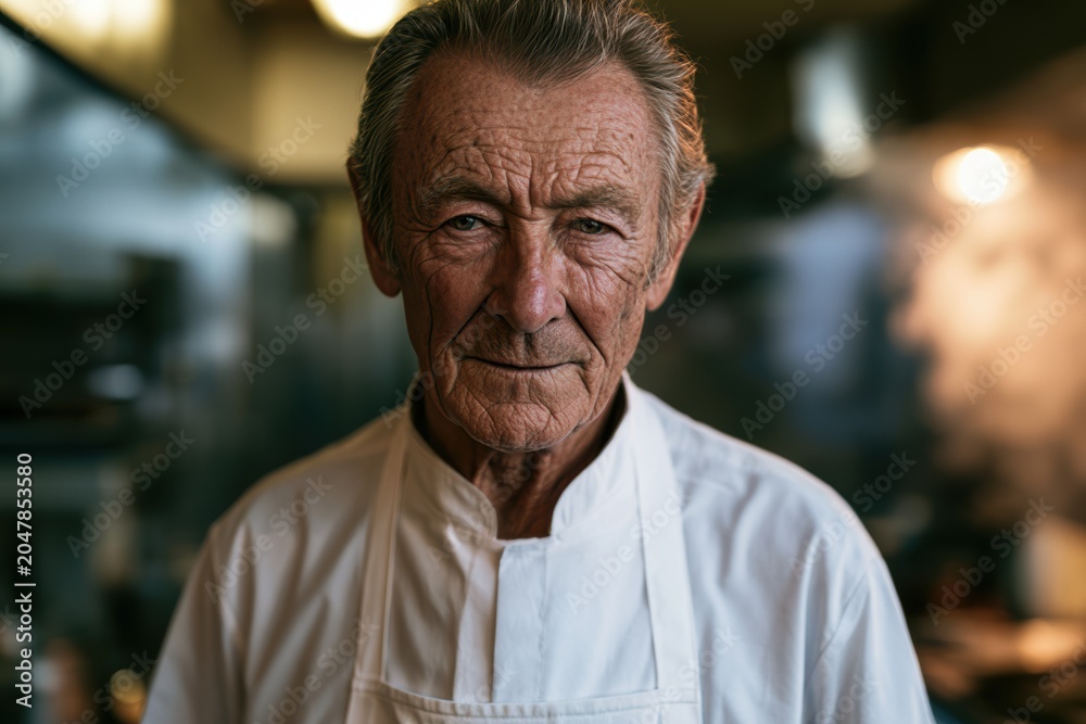 Fototapeta premium Senior male chef wearing white uniform standing in a professional kitchen, determined look