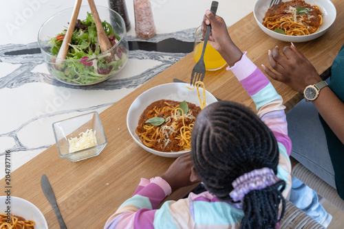 African American female mother and child eating spaghetti at wooden counter with salad, juice