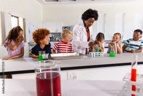African American teacher in white lab coat leading science demo in school lab students with goggles