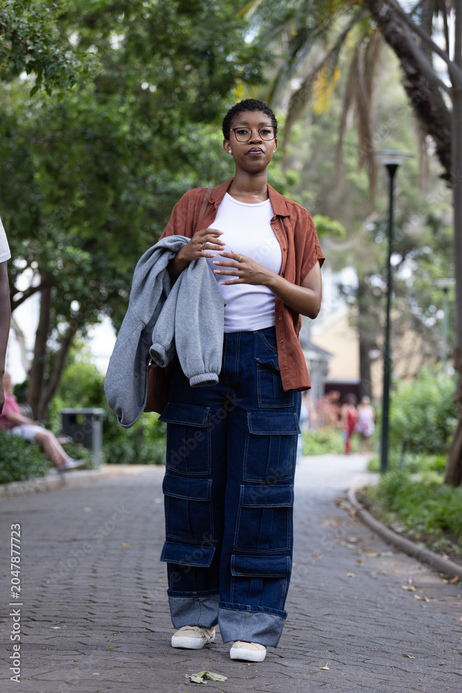 Fototapeta premium African woman walking on paved park path holding gray sweatshirt, wearing glasses and sneakers