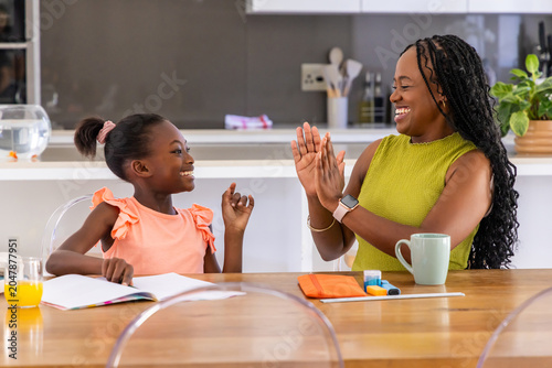African mother and youth daughter wearing lime and peach tops writing in notebook at kitchen table