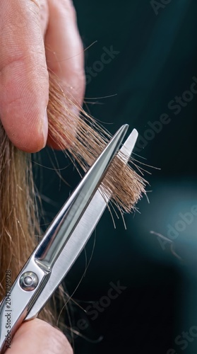 Macro view of fingers holding blonde hair ends while sharp metal scissors trim the strands against a dark blurred background.