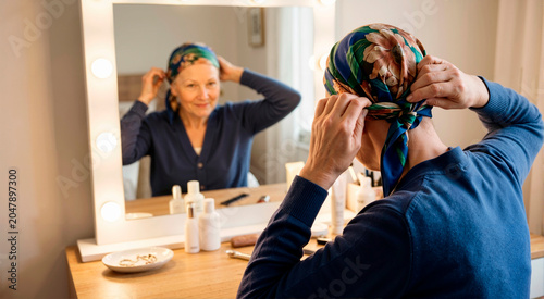 Senior woman tying a colorful headscarf in front of a vanity mirror. Elderly female cancer survivor smiling at her reflection. Hair loss and self-care concept