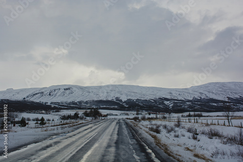 Wet asphalt road leading to snow covered mountains under overcast winter sky. High quality photo