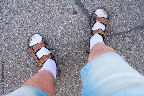 Man wearing white socks and brown sandals standing on asphalt pavement