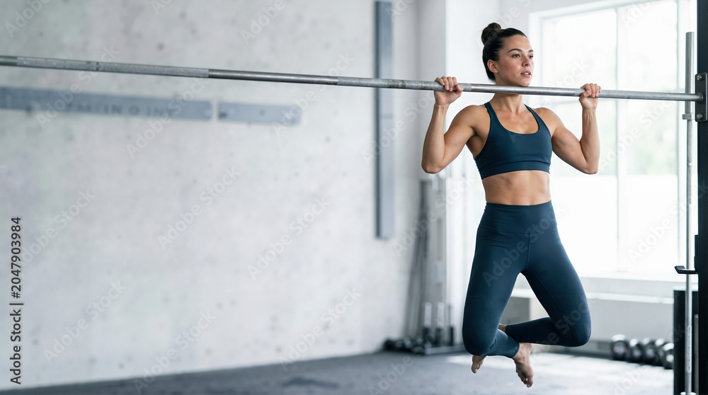 Fototapeta premium Female athlete performing a pull-up on a bar in a gym. Woman in sportswear doing upper body strength training workout. Fitness and calisthenics concept