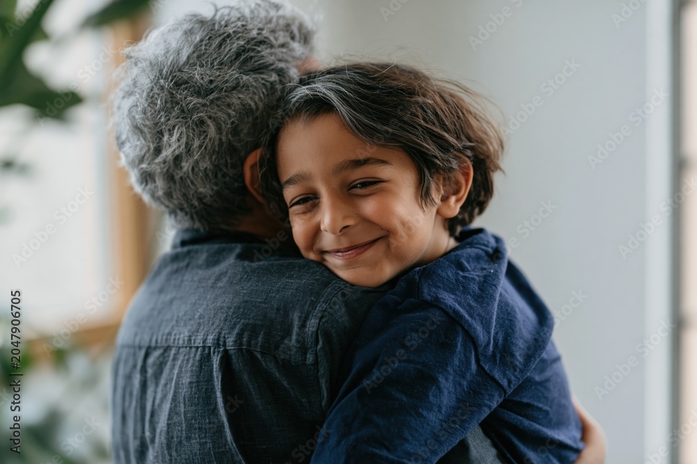 Obraz premium Smiling child hugged by older adult indoors for heartwarming family lifestyle portrait and comforting background
