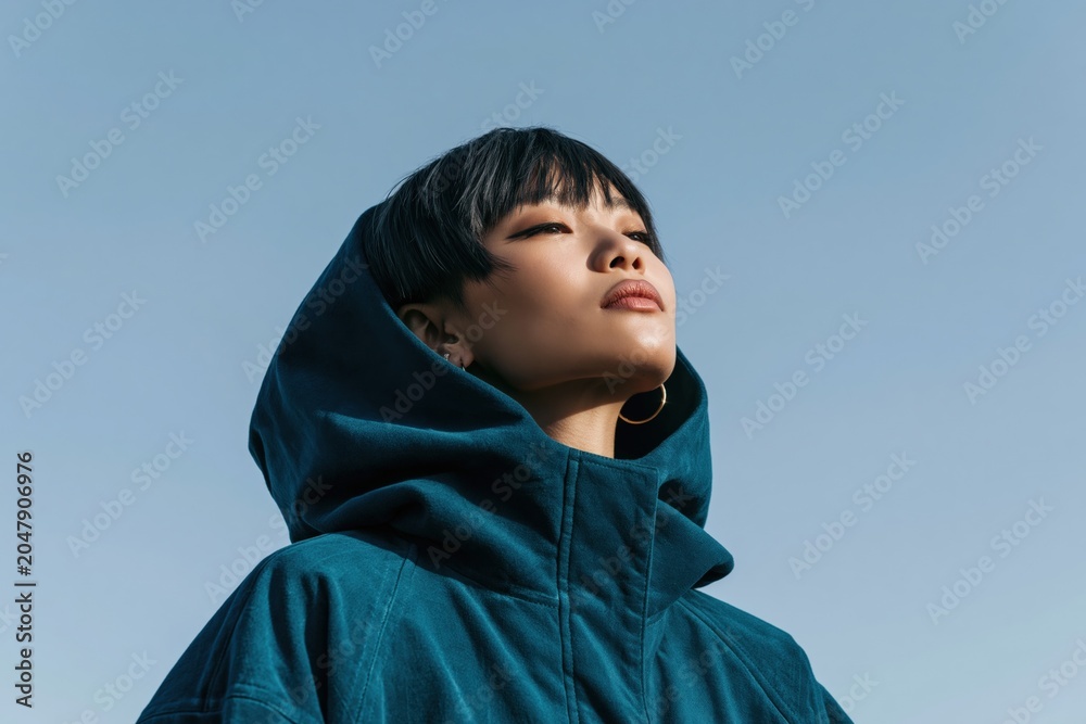 Obraz premium Young woman portrait in a teal hooded coat looking upward into clear sky for fashion banner and minimalist editorial background