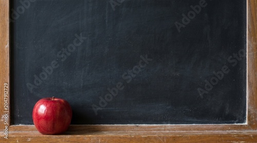 A single, ripe red apple sits on a wooden ledge in front of a blank blackboard, symbolizing learning and knowledge