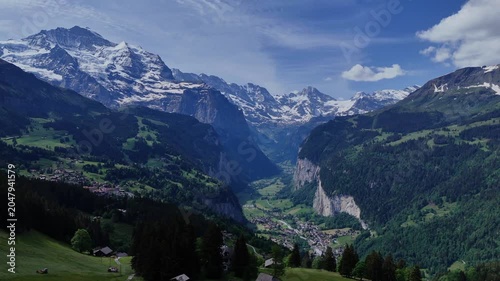 Sweeping drone panorama of Lauterbrunnen Valley, Swiss Alps, with alpine villages, steep cliffs, green slopes and snowcapped peaks under a blue sky.