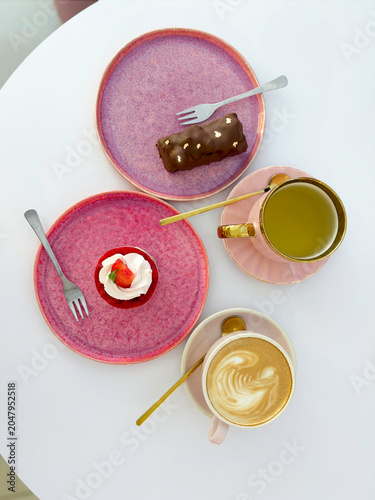 Vertical flat lay of coffee cup with latte art, green tea and mini cakes on pink plates. High quality photo