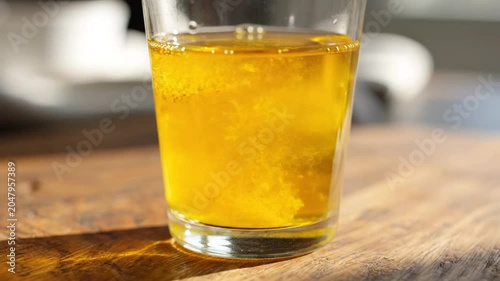 Pouring golden liquid into a glass, creating a swirling effect on a wooden table with sunlight.