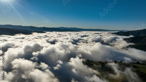 Breathtaking aerial view of fluffy clouds filling valley with distant mountains under clear blue sky during sunny day