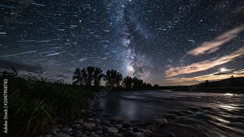 Time-lapse of starry night sky over flowing river with illuminated clouds and scenic landscape