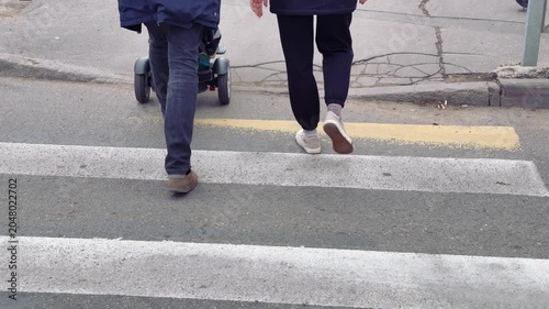 Couple pushing stroller crossing crosswalk, rear view of man and woman in casual jackets walking on worn zebra stripes, stroller