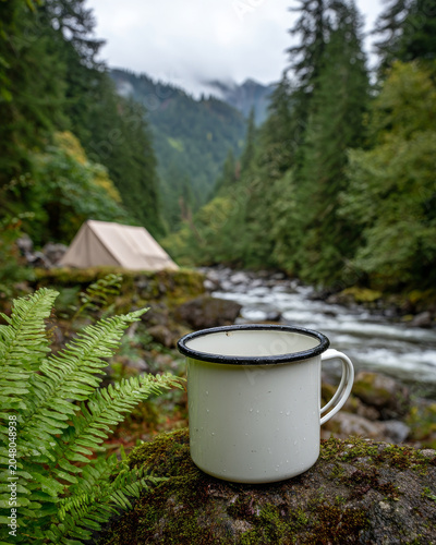White enamel camping mug with black rim sits on mossy boulder near flowing river and green forest with tent in background