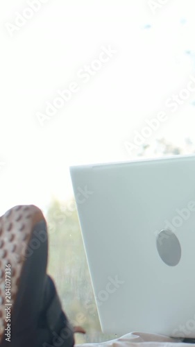 Vertical video: Shifting leg, person in joggers nudging laptop by sunlit window, black slide sandal