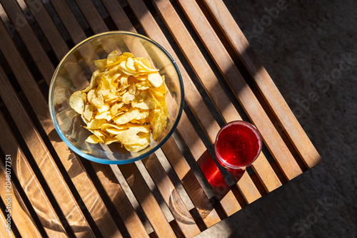 Clear glass bowl potato chips sitting on patio slat table beside short red tumbler casting stripes