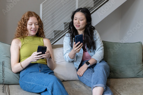 Quadro su tela Diverse female friends sitting on sofa in living room comparing smartphones with